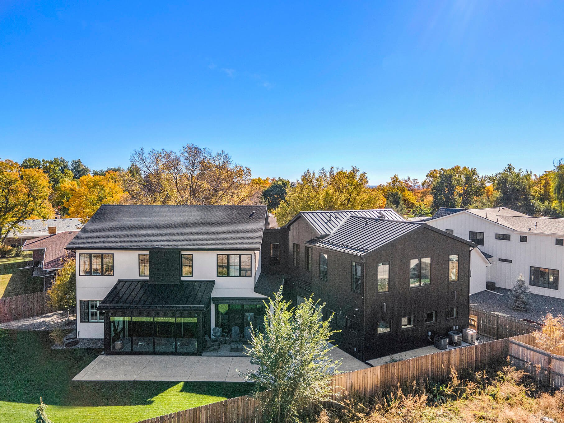 Aerial view of a modern suburban house with a large backyard, surrounded by autumn trees. The house features a mix of white and dark exterior finishes, a black roof, and a paved patio area. Neighboring houses and a wooden fence are visible.