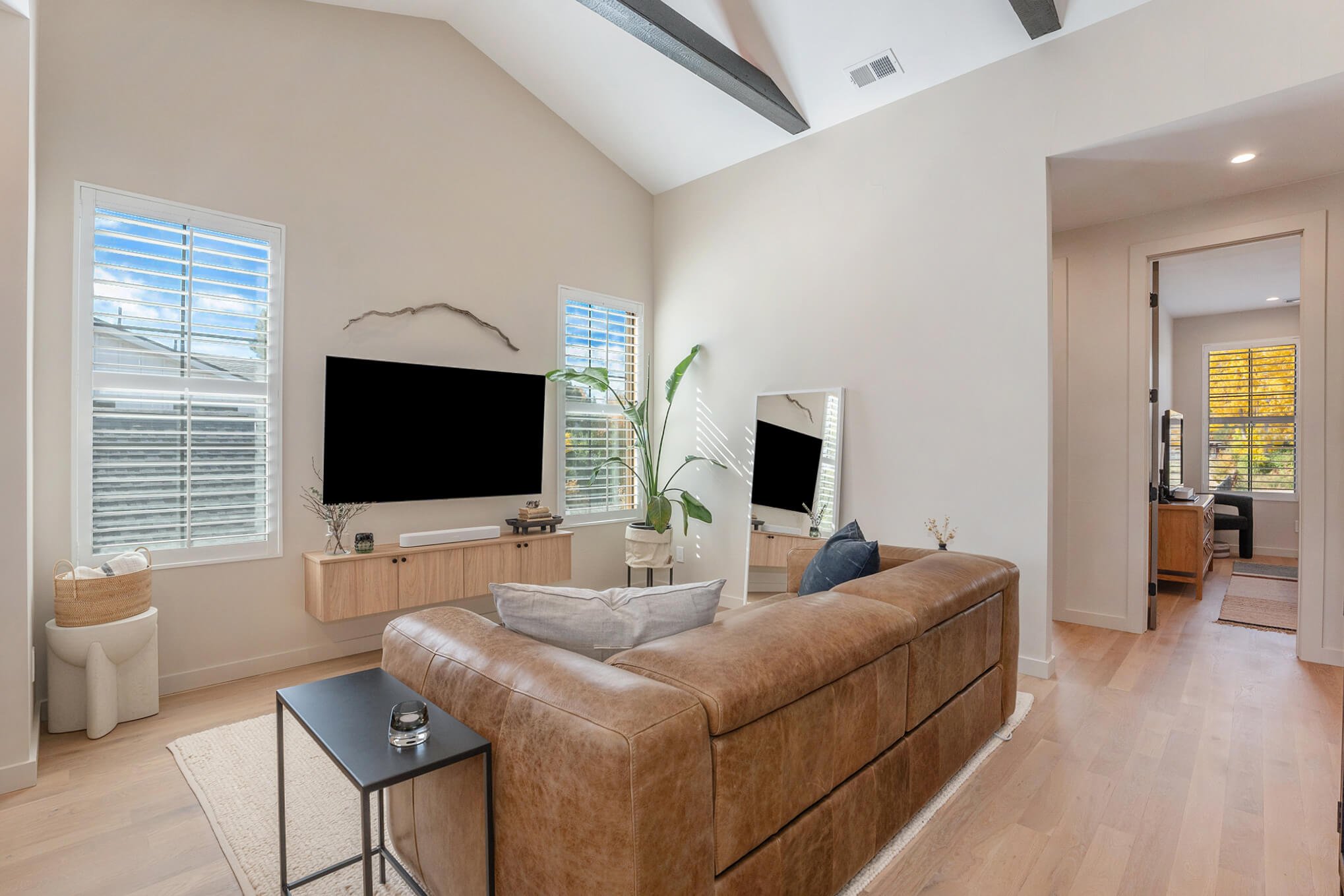 Modern living room with a brown leather sofa, wall-mounted TV, wooden console, large mirror, tall indoor plant, light rug, and light wood flooring. Adjacent room visible through an open doorway.