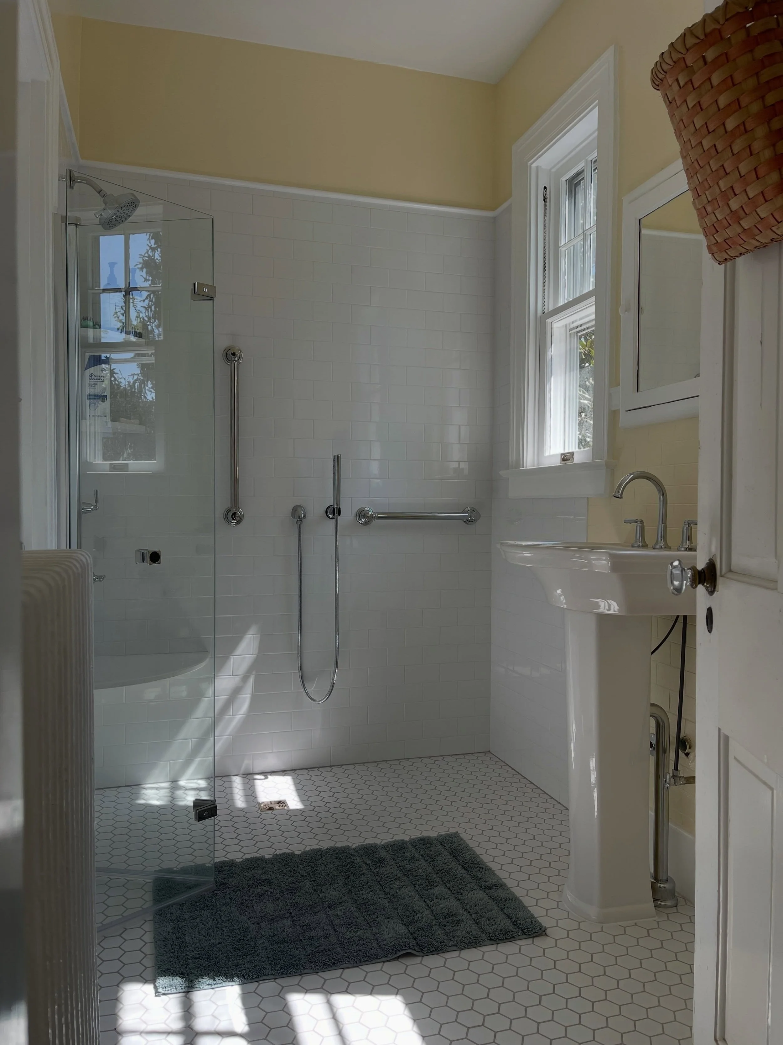 Our latest bathroom renovation in a storybook historic home in Downtown Winchester features a curb-less shower with a bi-fold glass door for seamless access. Stamped lime plaster in a running bond pattern matches the original walls. The bucketfuls of