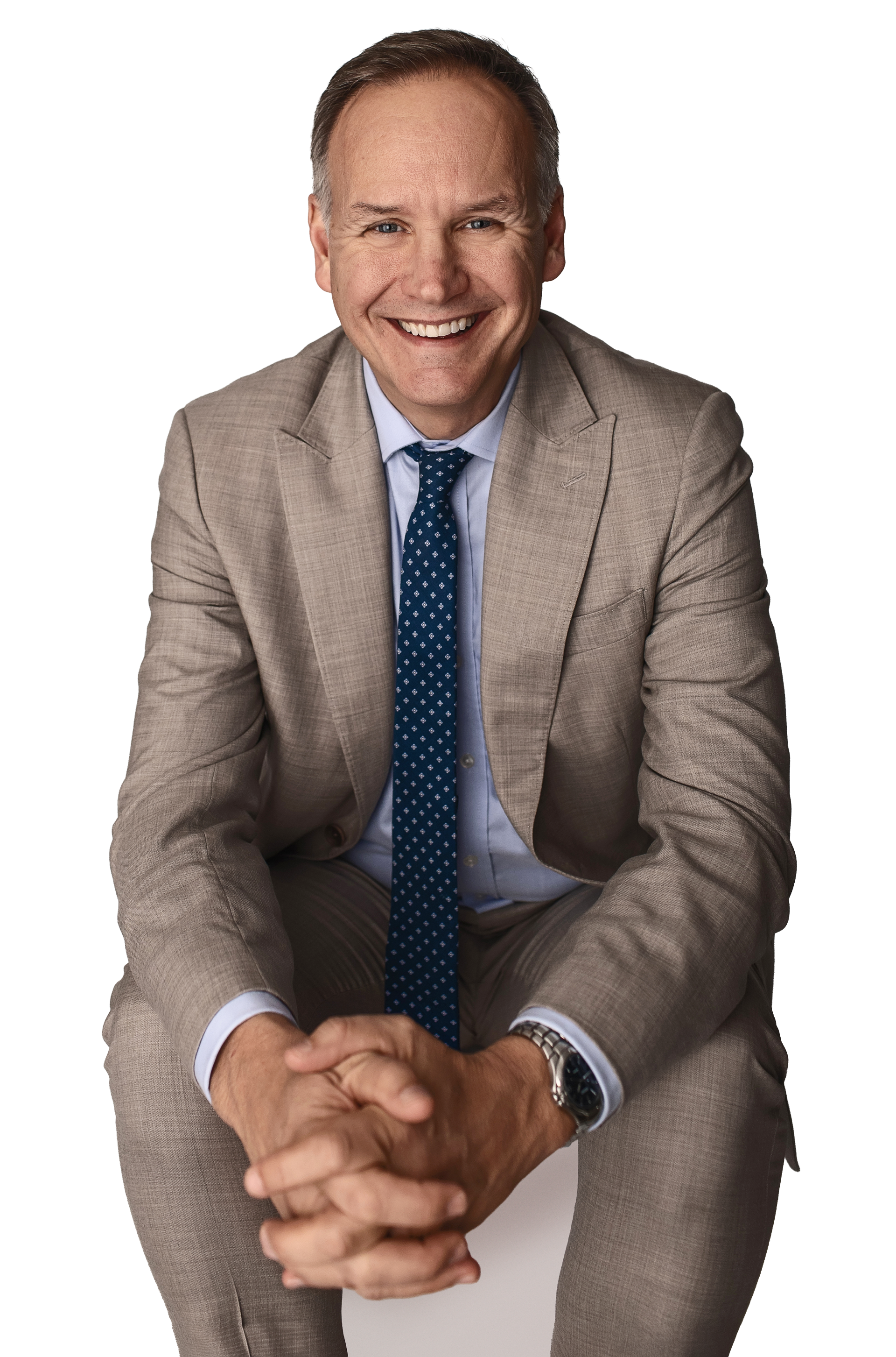 A middle-aged man with a friendly smile, wearing a beige suit, light blue shirt, dark blue patterned tie, and a wristwatch, sitting with his hands clasped together.