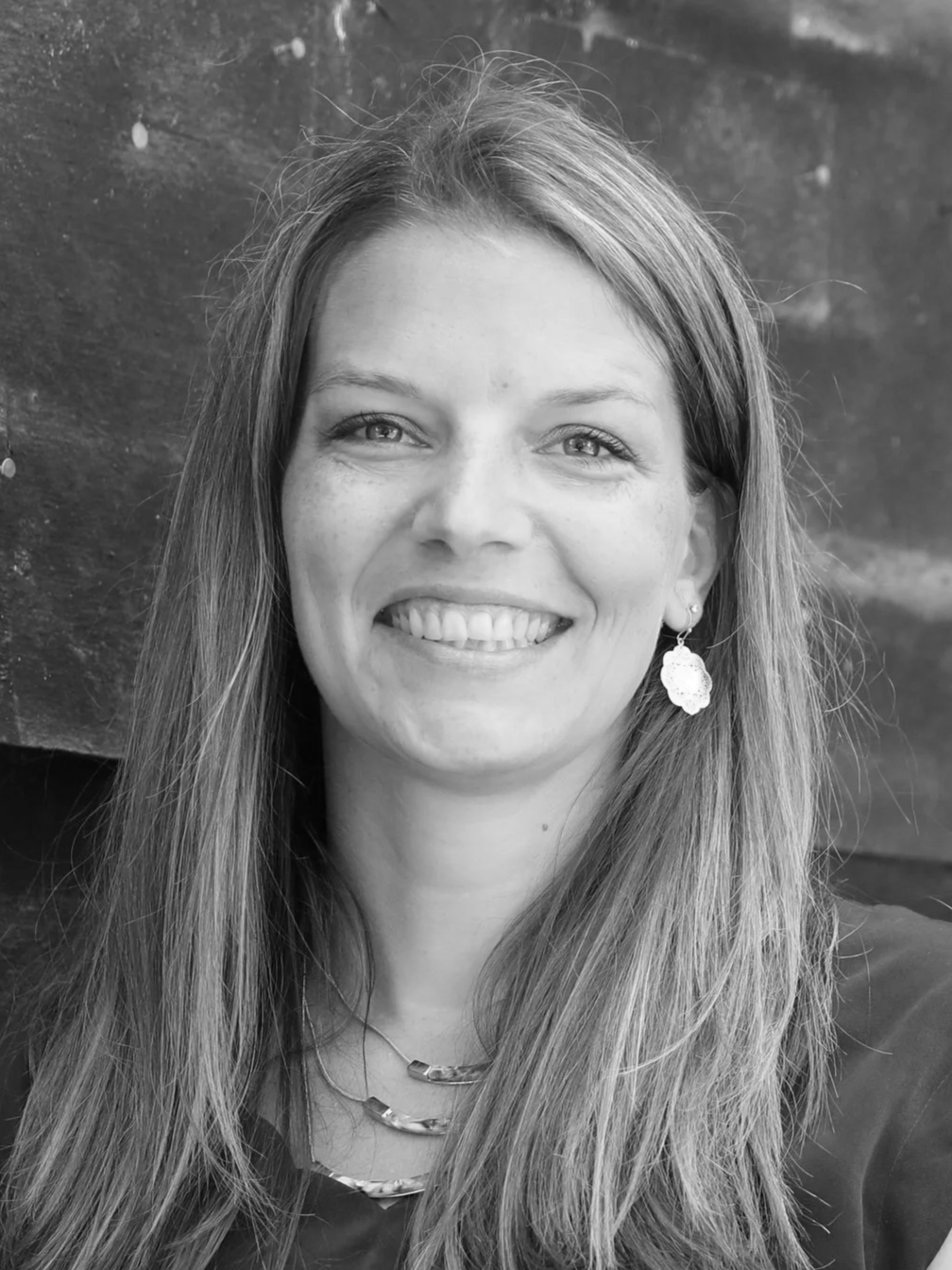A smiling woman with long hair wearing earrings and a necklace, posing outdoors against a wooden background.