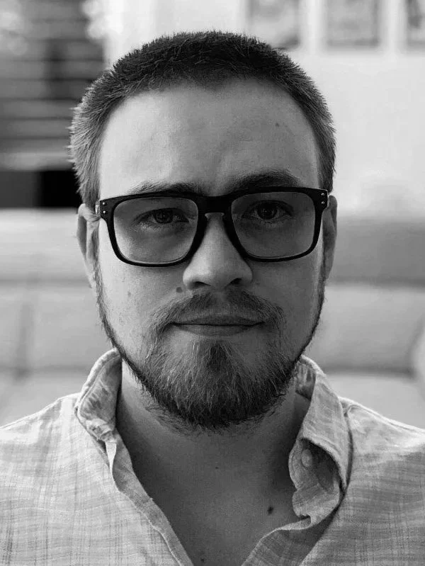 A close-up black-and-white photo of a young man with short hair, glasses, a beard, wearing a collared shirt, looking directly at the camera.