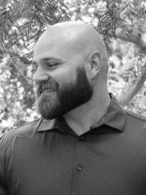 A black and white photo of a man with a shaved head and full beard, smiling and looking to his left, outdoors with trees in the background, wearing a collared shirt.
