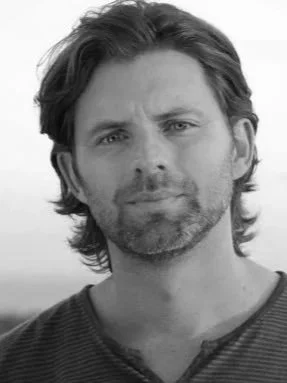 Black and white portrait of a man with shoulder-length wavy hair and a beard, wearing a striped shirt, looking at the camera with a neutral expression.