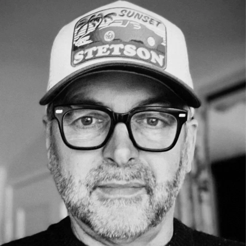 A man wearing glasses and a cap that says 'Sunset Stetson' in a close-up shot, black and white photo.