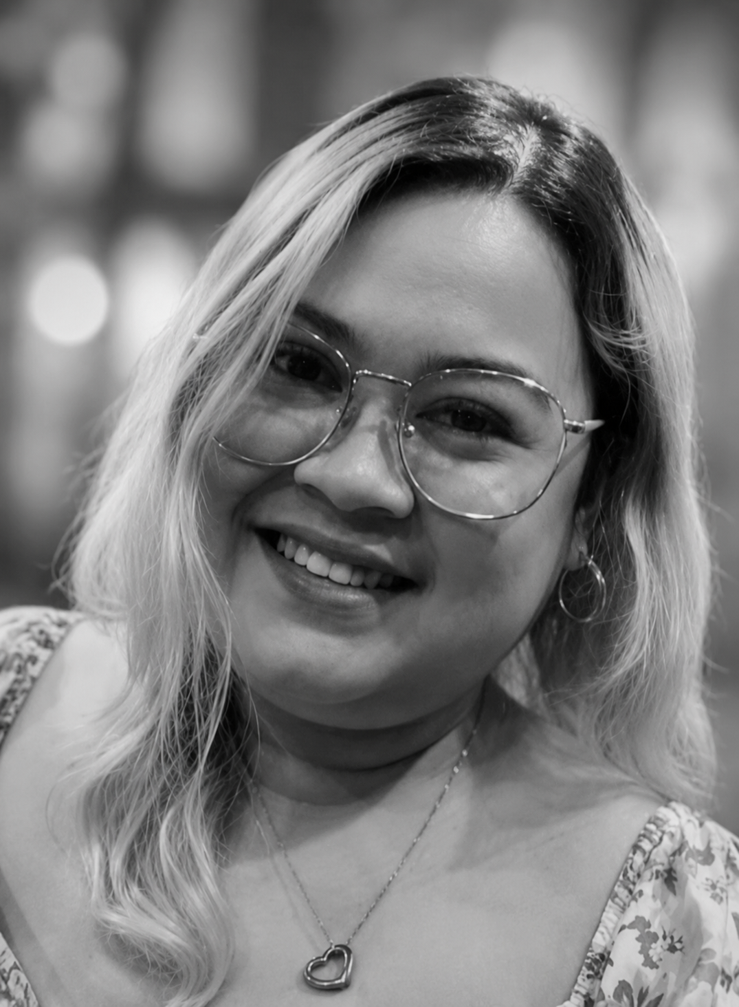 A woman smiling, wearing glasses, jewelry, and a patterned top, in a black and white photo.