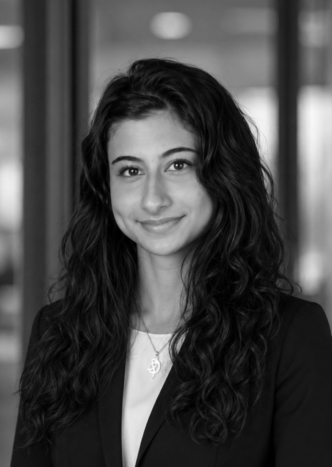 Black and white portrait of a young woman with long curly hair, wearing a blazer and a necklace, smiling at the camera.