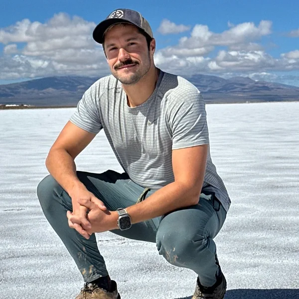 A man with a beard and mustache, wearing a gray t-shirt, gray pants, a black wristwatch, and a black cap, squatting on a salt flat with mountains and a partly cloudy sky in the background.