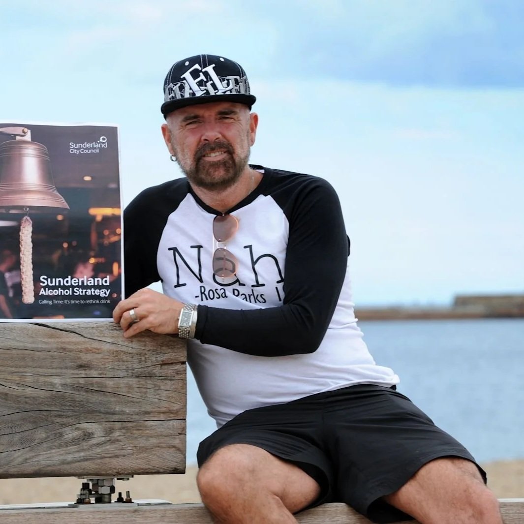 A man with a beard wearing a black and white baseball cap, sunglasses hanging from his shirt, and a shirt that says "Noh Rosa Parks". He is sitting on a wooden bench near water, holding a sign that promotes Sunderland's alcohol strategy.