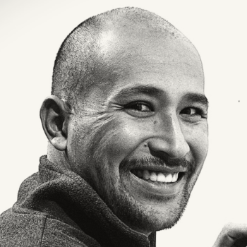 Black and white portrait of a smiling man with a shaved head, looking over his shoulder.