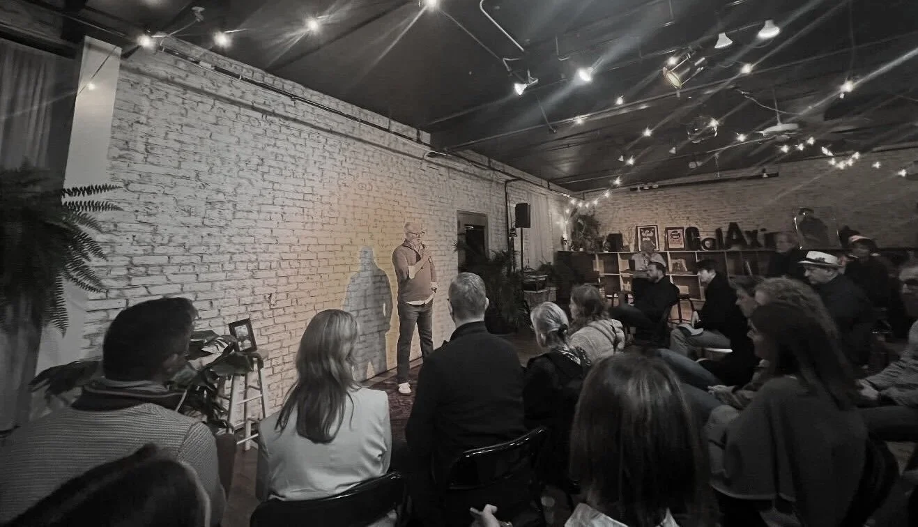 A man standing with a microphone speaking to an audience in a dimly lit room with string lights, a white brick wall, and shelves in the background.