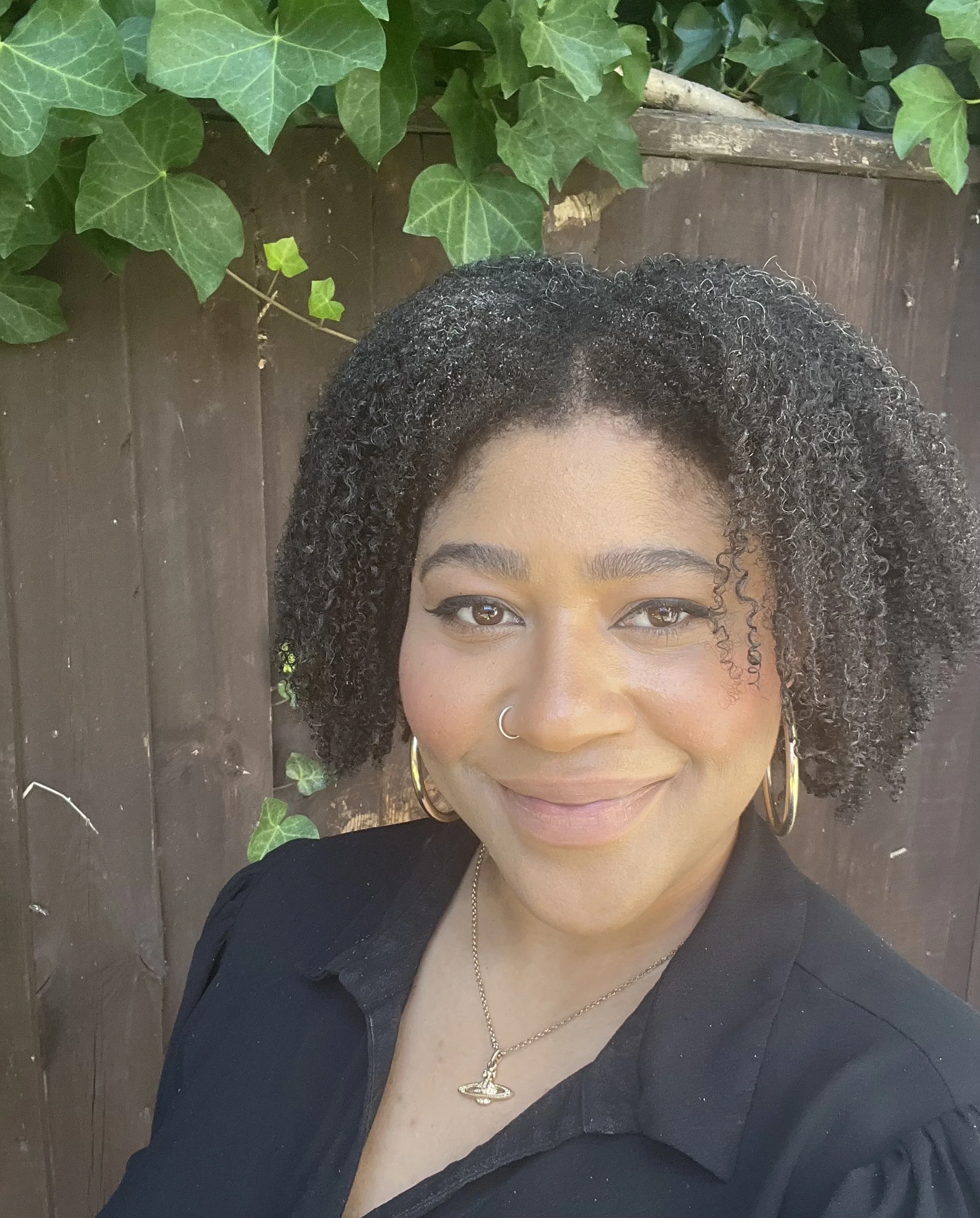 A woman with curly hair, brown eyes, and a nose piercing, smiling, wearing gold hoop earrings, a gold necklace, and a black top, standing in front of a wooden fence with green leafy ivy plants.