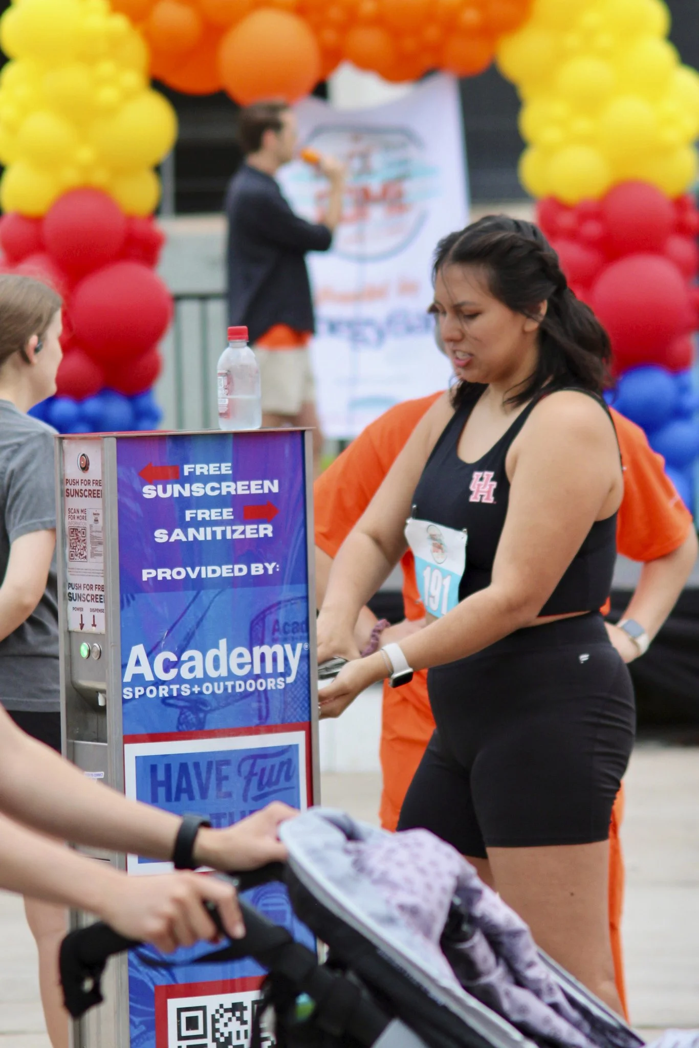 A woman in a black athletic outfit receives sunscreen and sanitizer from a station at a sports event, with balloons in red, yellow, and blue colors in the background.