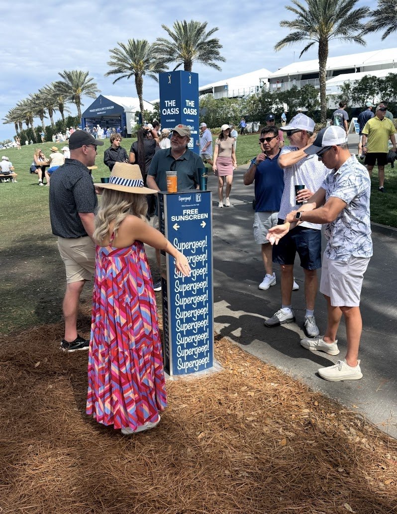 People surrounding a Supergoop-branded sunscreen dispenser at THE PLAYERS Championship providing free sunscreen for fans at major outdoor sporting events.