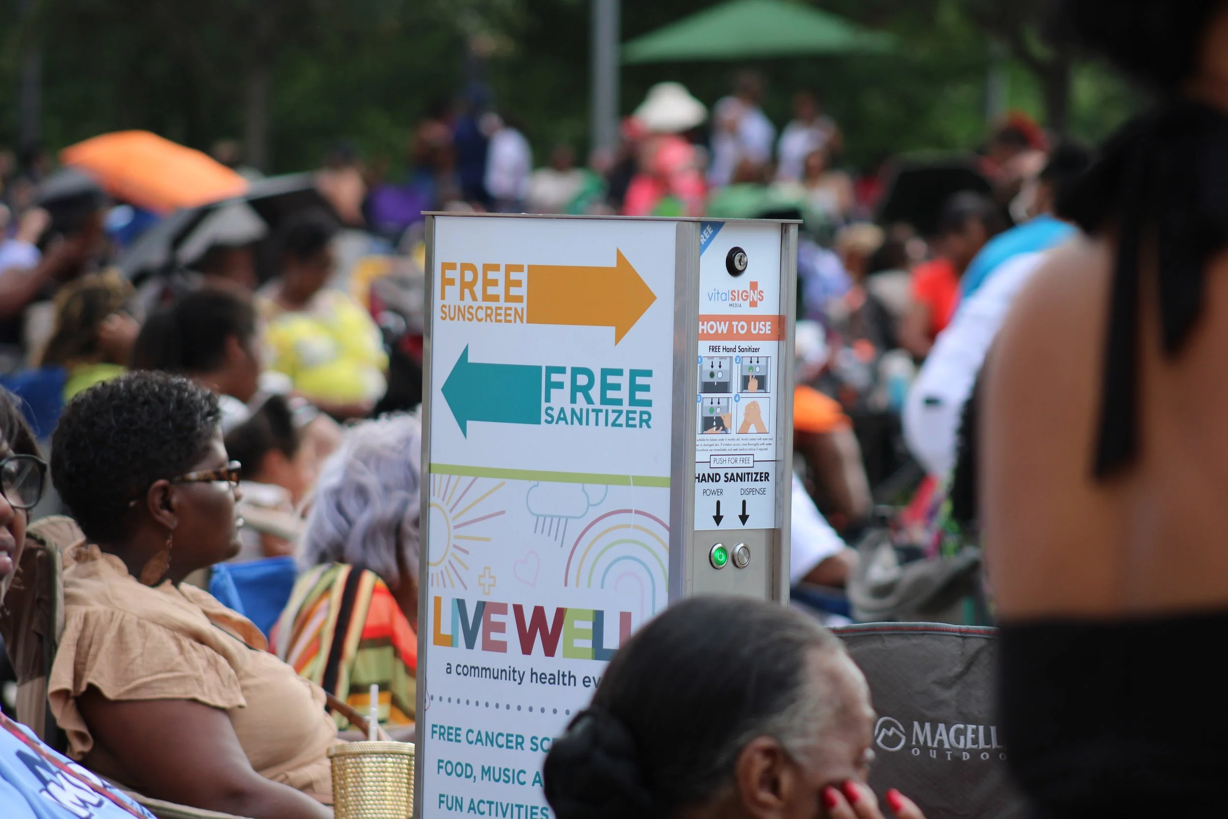 A crowd of people sitting outdoors at a community health event. A kiosk with free sunscreen to the right and free sanitizer to the left, with instructions on how to use.