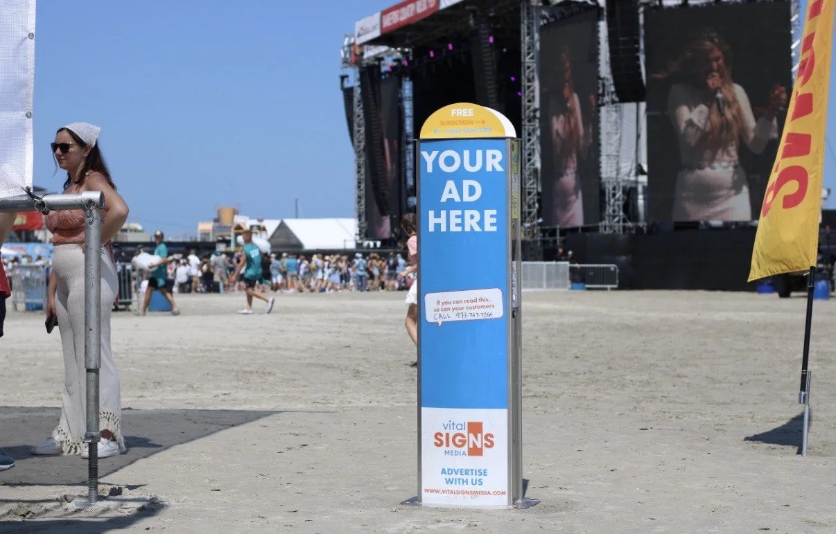 A promotional sign on a beach with a stage and large video screens in the background during a sunny day. The sunscreen kiosk has a placeholder for an advertisement.