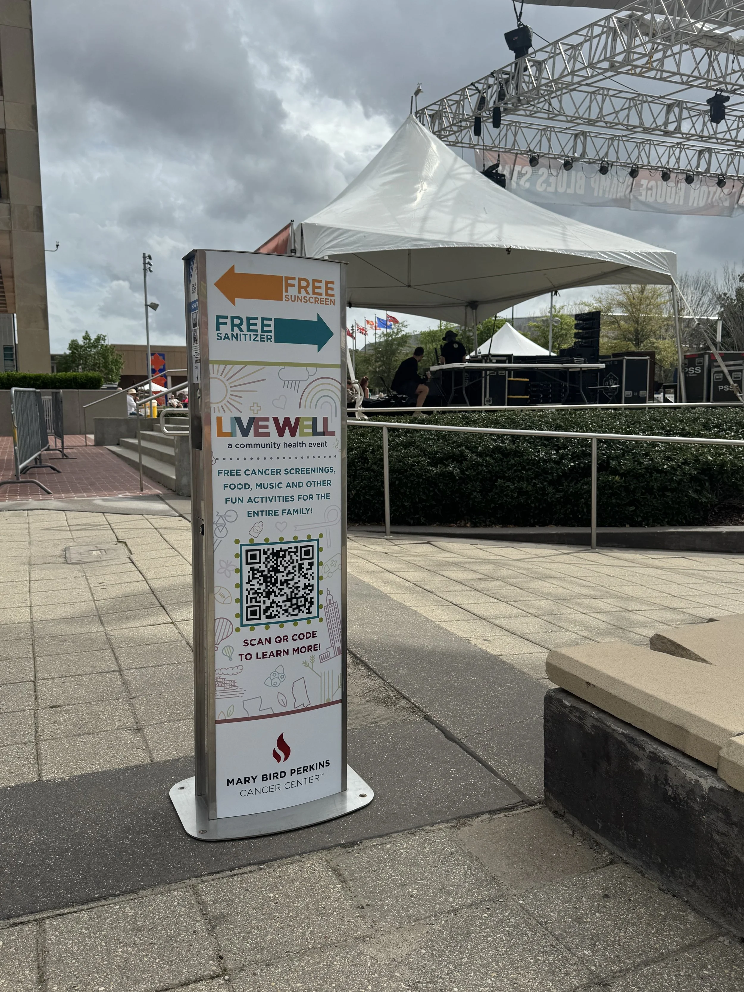 Vital Signs Media sunscreen kiosk promoting a community health event with free sunscreen, free sanitizer, QR code, and event details, located outdoors near a stage with performers under a white canopy, with cloudy sky overhead.