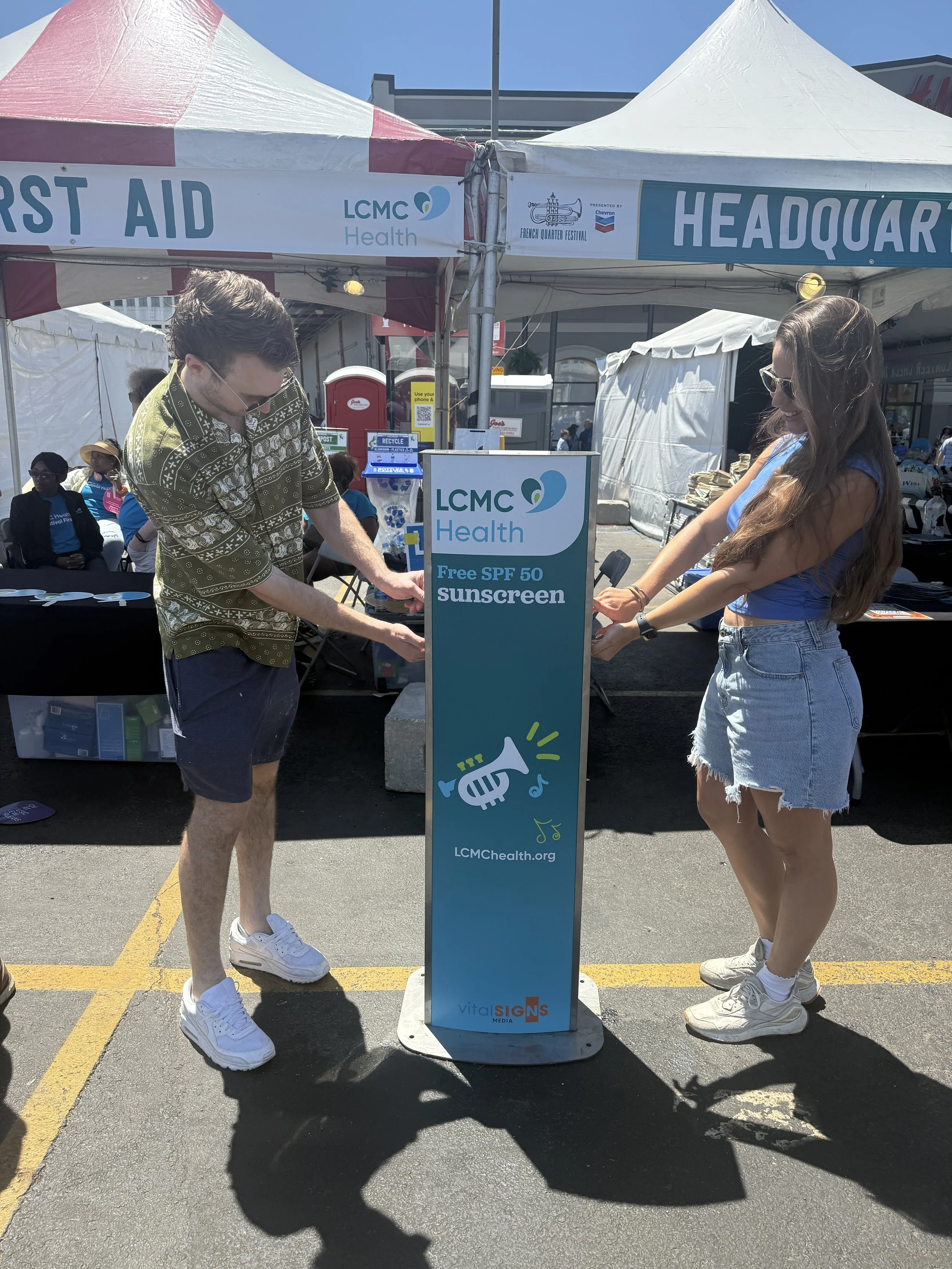 A man and woman putting on sunscreen next to a Vital Signs Media sunscreen dispenser. The dispenser reads, "LCMC Health Free SPF 50 sunscreen" and features the LCMC Health logo and website.