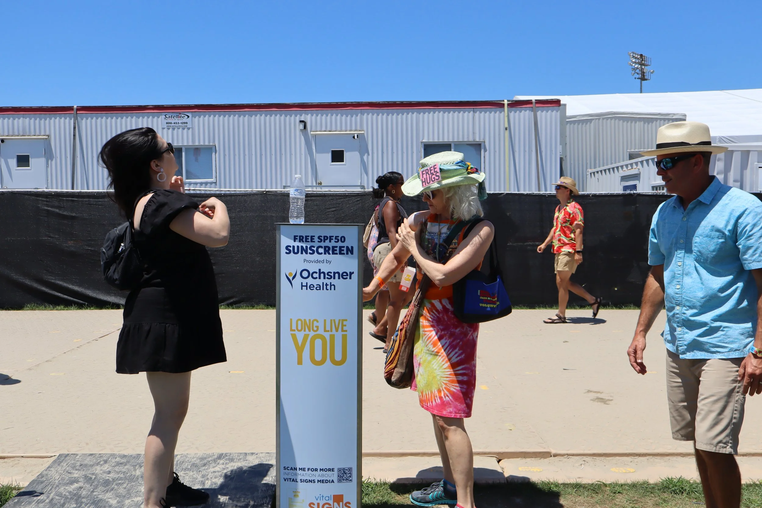 A woman in a black dress and sunglasses is applying sunscreen at a free SPF50 sunscreen station, with other people walking in the background on a sunny day.