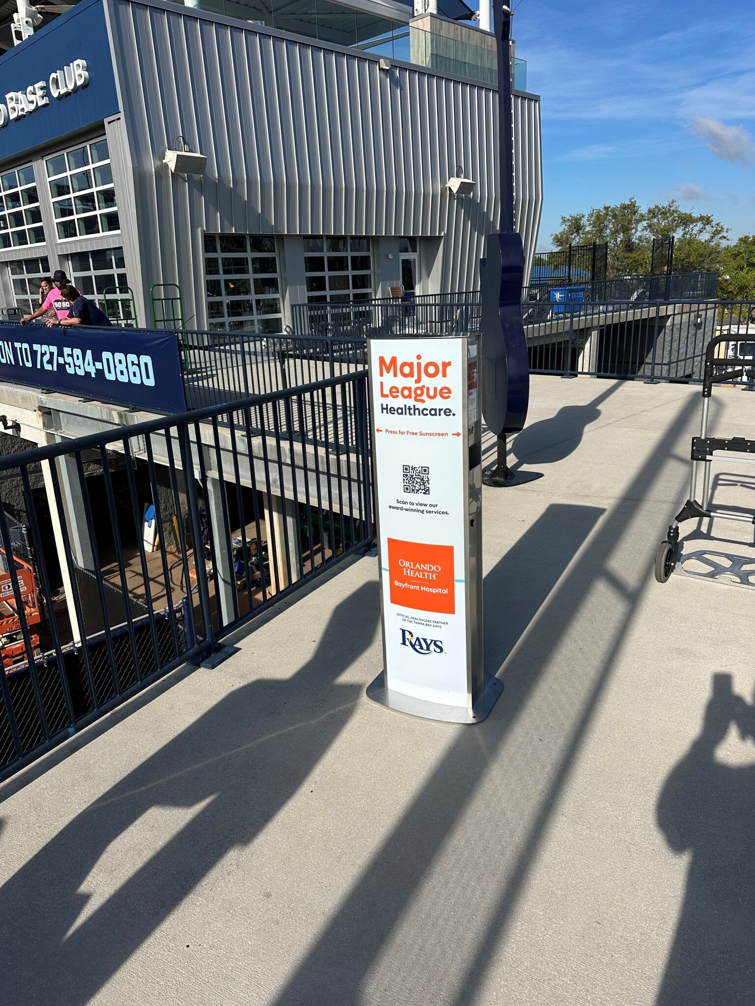 Sunscreen kiosk outside at the Tampa Bay Rays stadium sponsored by Major League Healthcare at Orlando Health, Bayfront Hospital, with QR code and contact information, on a concrete sidewalk with shadows and nearby railing.