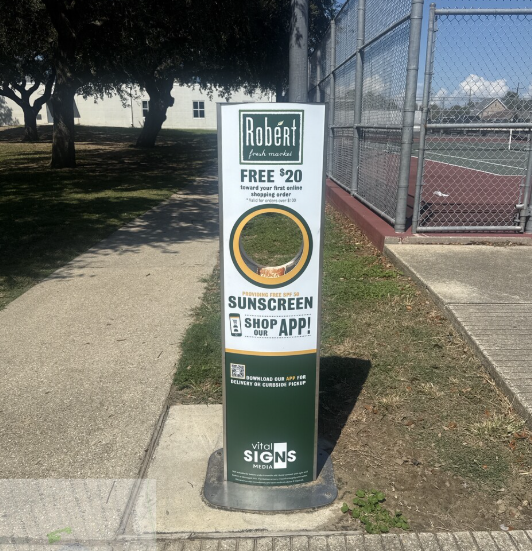 Kiosk advertising a free $20 online shopping credit at Robert Fresh Market, dispensing free sunscreen app, located next to a tennis court on a sunny day.