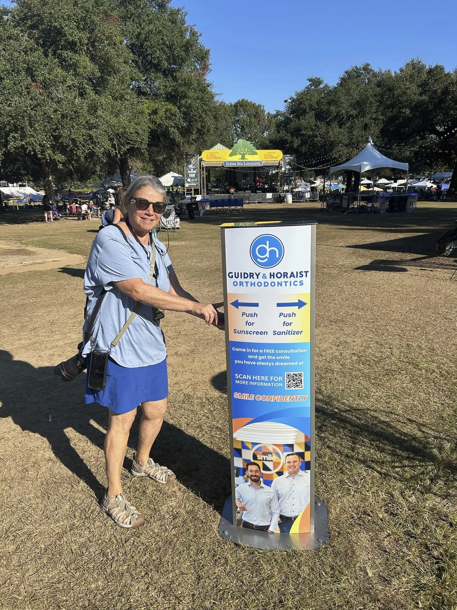 Woman wearing sunglasses getting hand sanitizer out of a sunscreen kiosk at an outdoor event or festival.