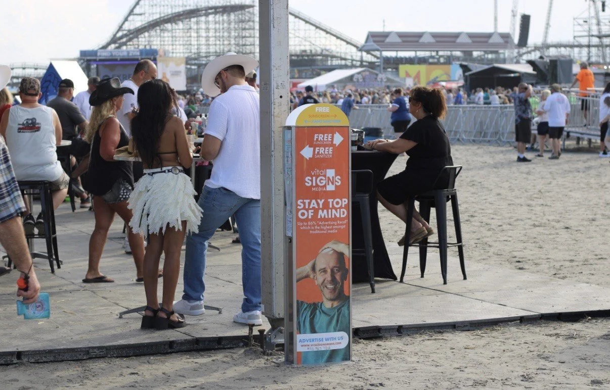 People on a beach boardwalk near an amusement park, with a woman sitting at a table and a kiosk offeirng free sunscreen and sanitizer.