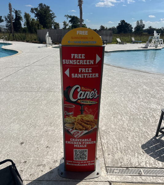 A red and yellow kiosk near a swimming pool offers free sunscreen and hand sanitizer, and promotes Raising Cane's chicken finger meals with an image of fried chicken fingers, fries, and dipping sauces.