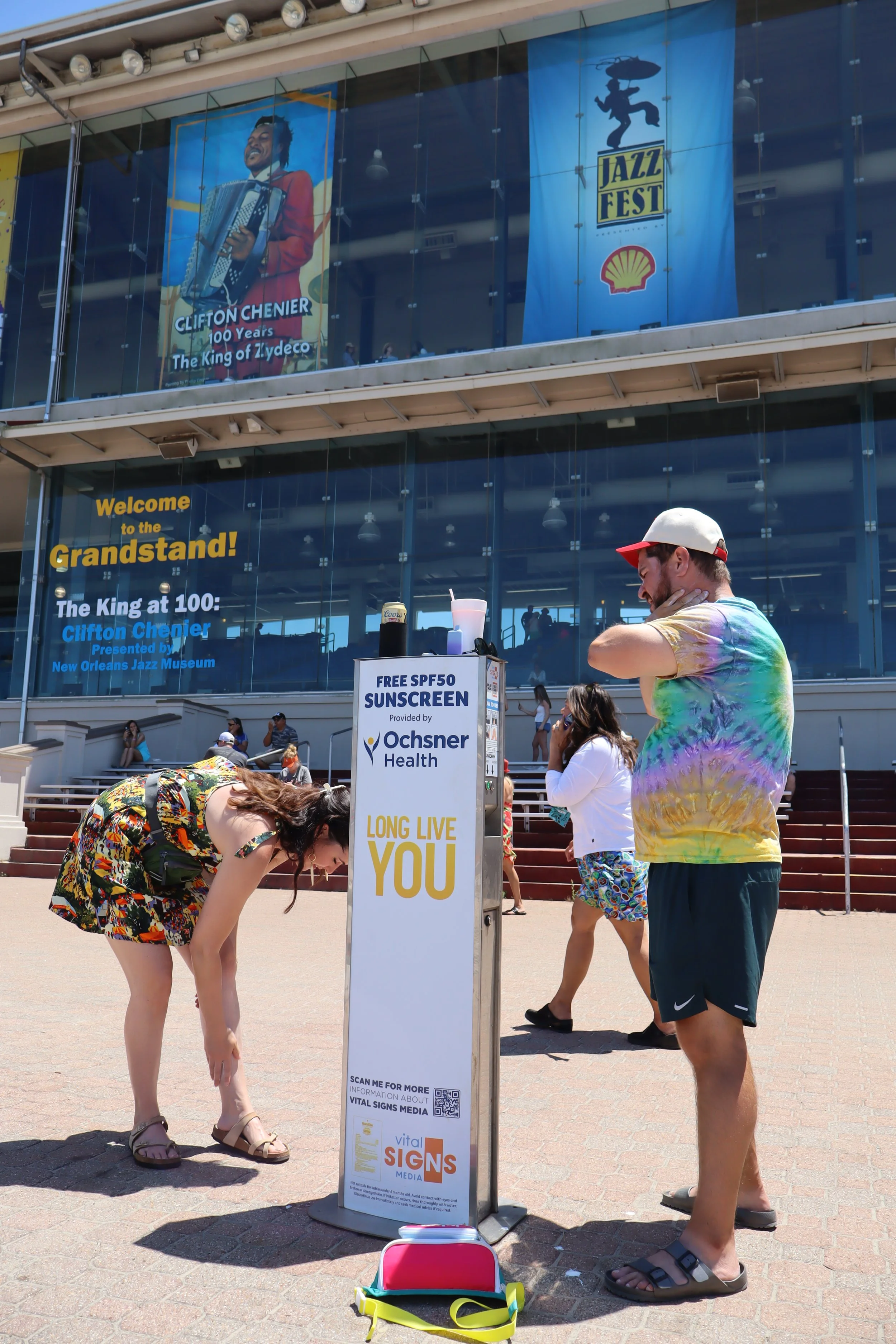 People standing outdoors near a kiosk offering free SPF50 sunscreen at a jazz festival. The festival celebrates Clifton Chenier's 100th birthday with banners and a glass-front building with steps leading up to it.