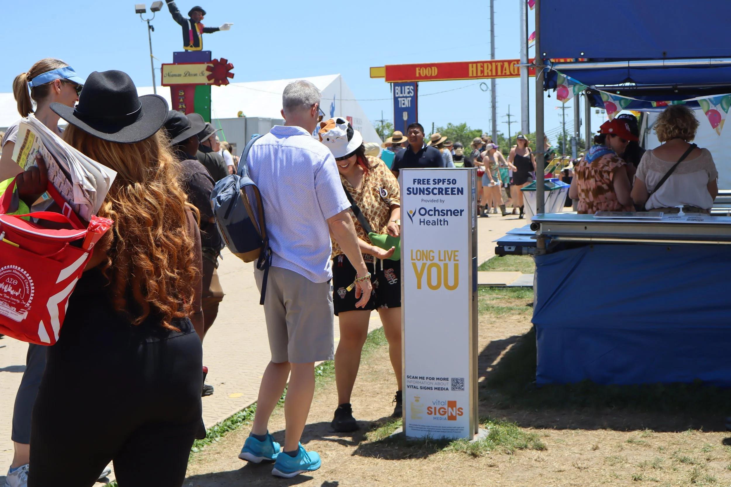 Crowd at outdoor fair or festival with people lined up at a Vital Signs Media kiosk offering free SPF50 sunscreen. There is signage from Ochsner Health and a woman in the line is smiling and interacting with the staff at the booth.