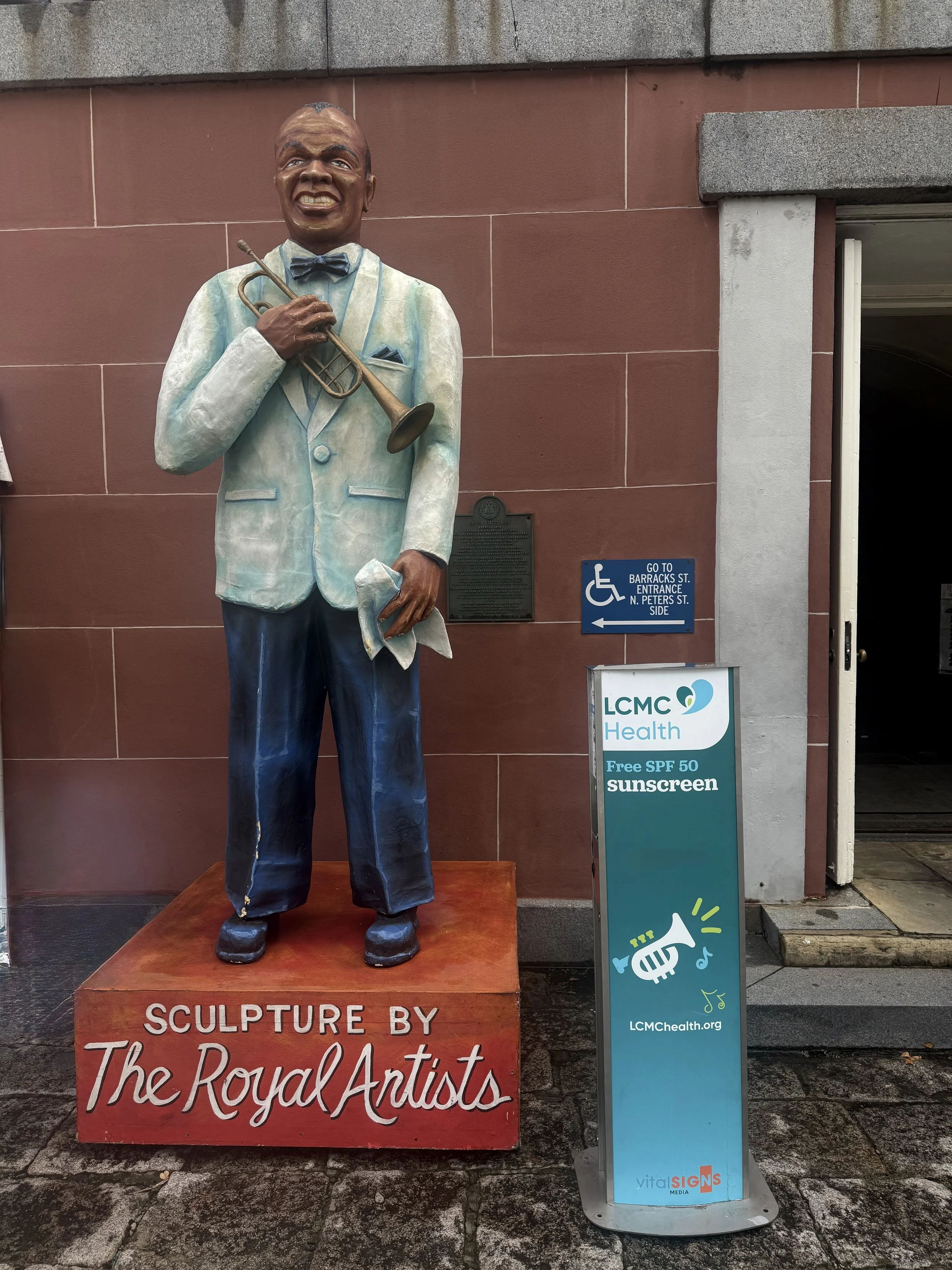 A colorful statue of a man in a light suit holding a trumpet and a cloth, standing on a red pedestal labeled 'Sculpture by The Royal Antistas,' outside a building next to a sunscreen dispenser offering free SPF 50 sunscreen.