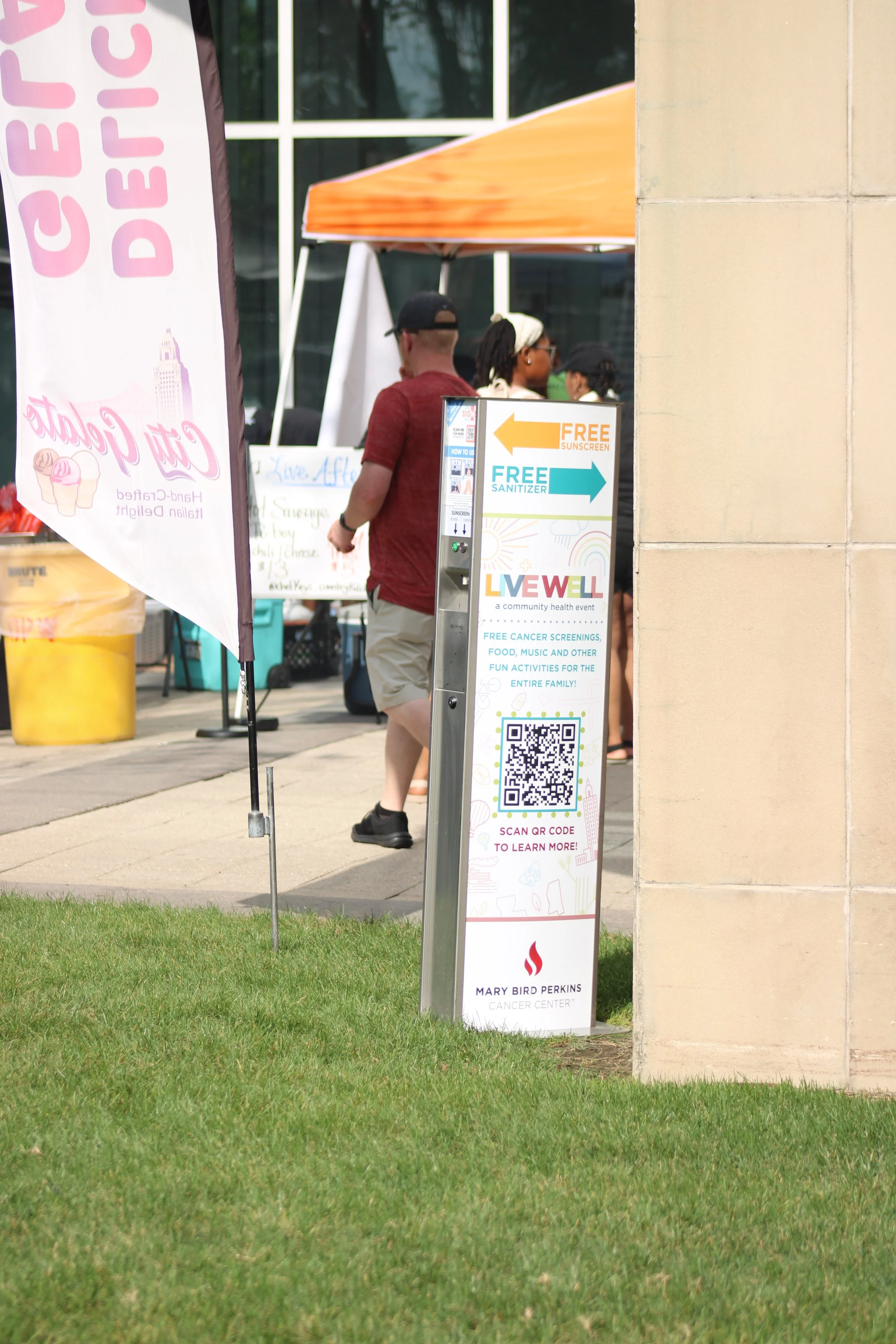 Vital Signs Media sunscreen dispenser with QR code for more information, in front of a building with people gathered nearby.