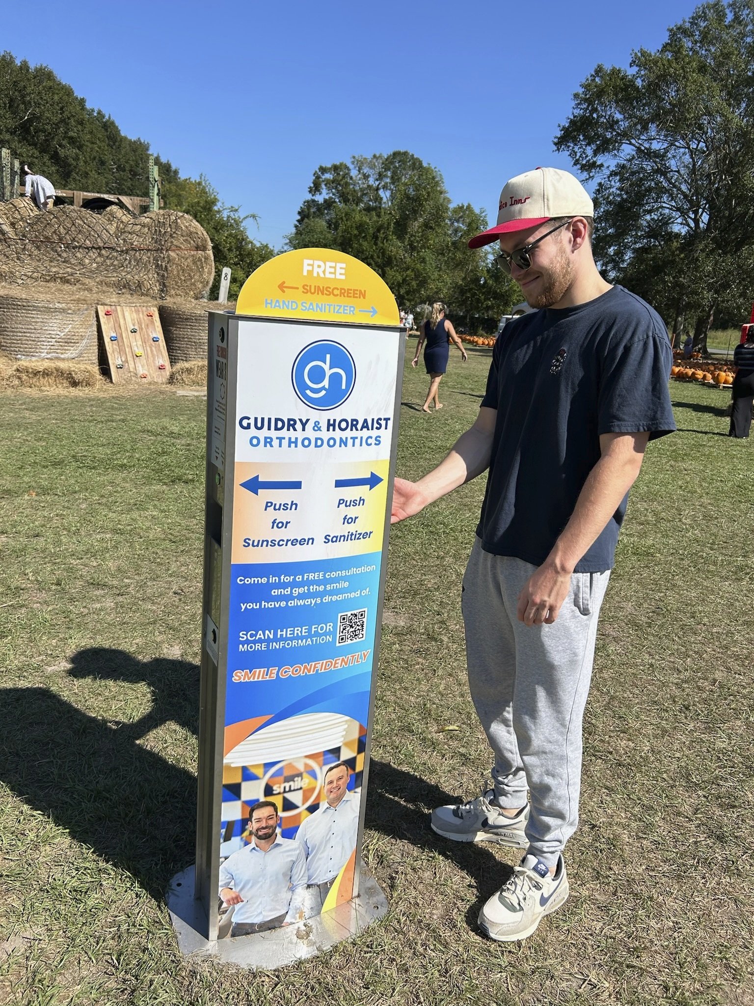 A man using a stand with a kiosk for Guidry & Horaist Orthodontics offering free sunscreen and hand sanitizer at an outdoor event, with hay bales and a climbing wall in the background.