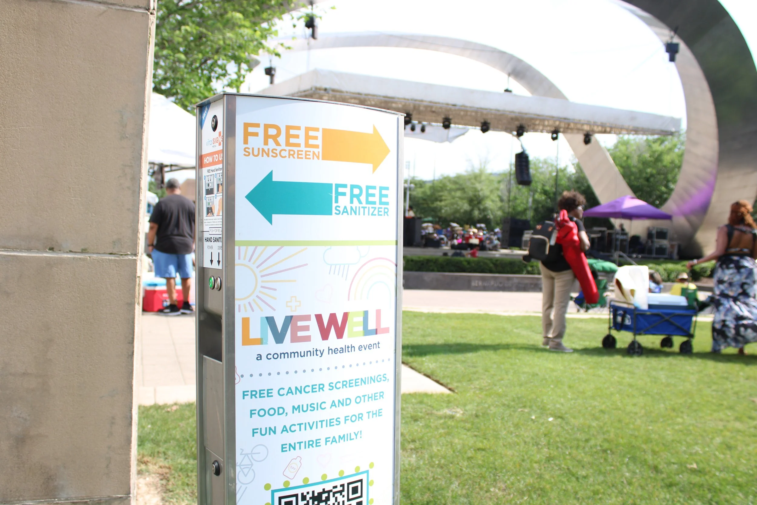 Free sunscreen and sanitizer station at a community health event called Live Well. In the background, people are gathering near a stage with musical performance, and some are pushing carts or wearing backpacks.