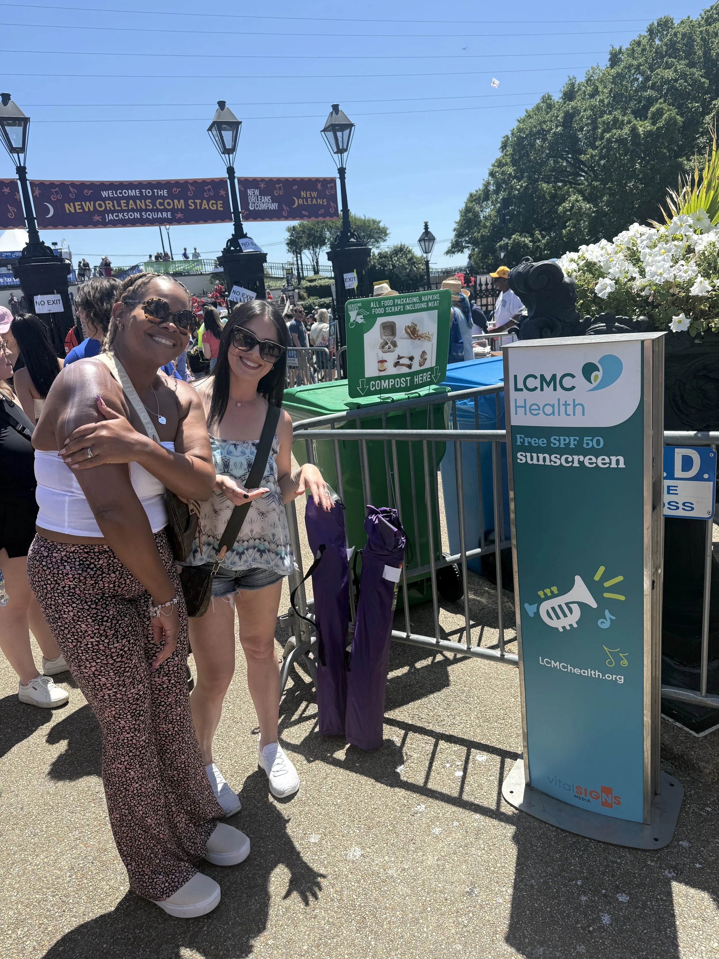 Two women smiling and posing at an outdoor event near a kiosk sponsored by LCMC Health offering free SPF 50 sunscreen, with a crowd, banners, and trees in the background.