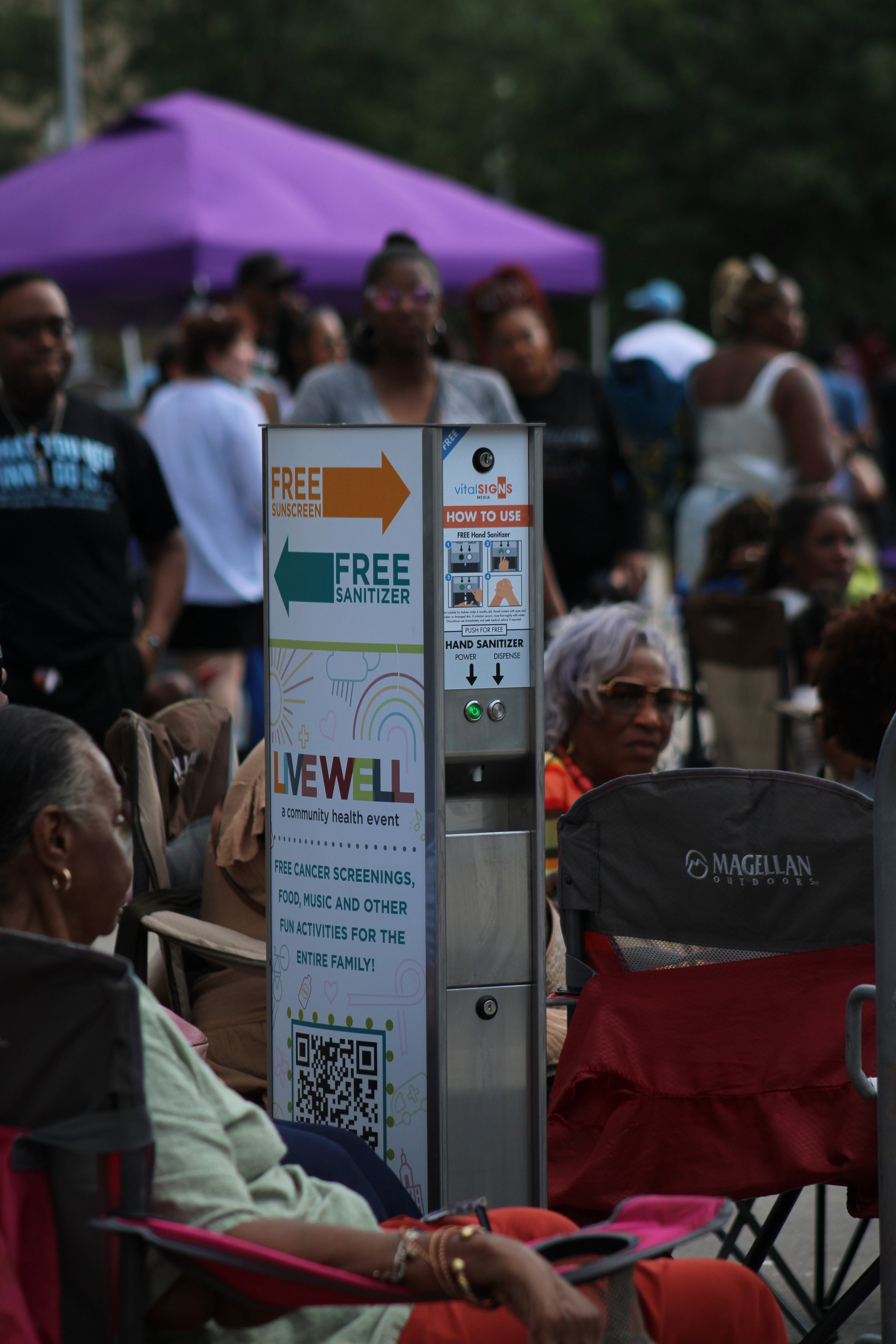 A community health event with people gathered around a sunscreen dispenser offering free sunscreen and sanitizer, with a purple tent in the background.