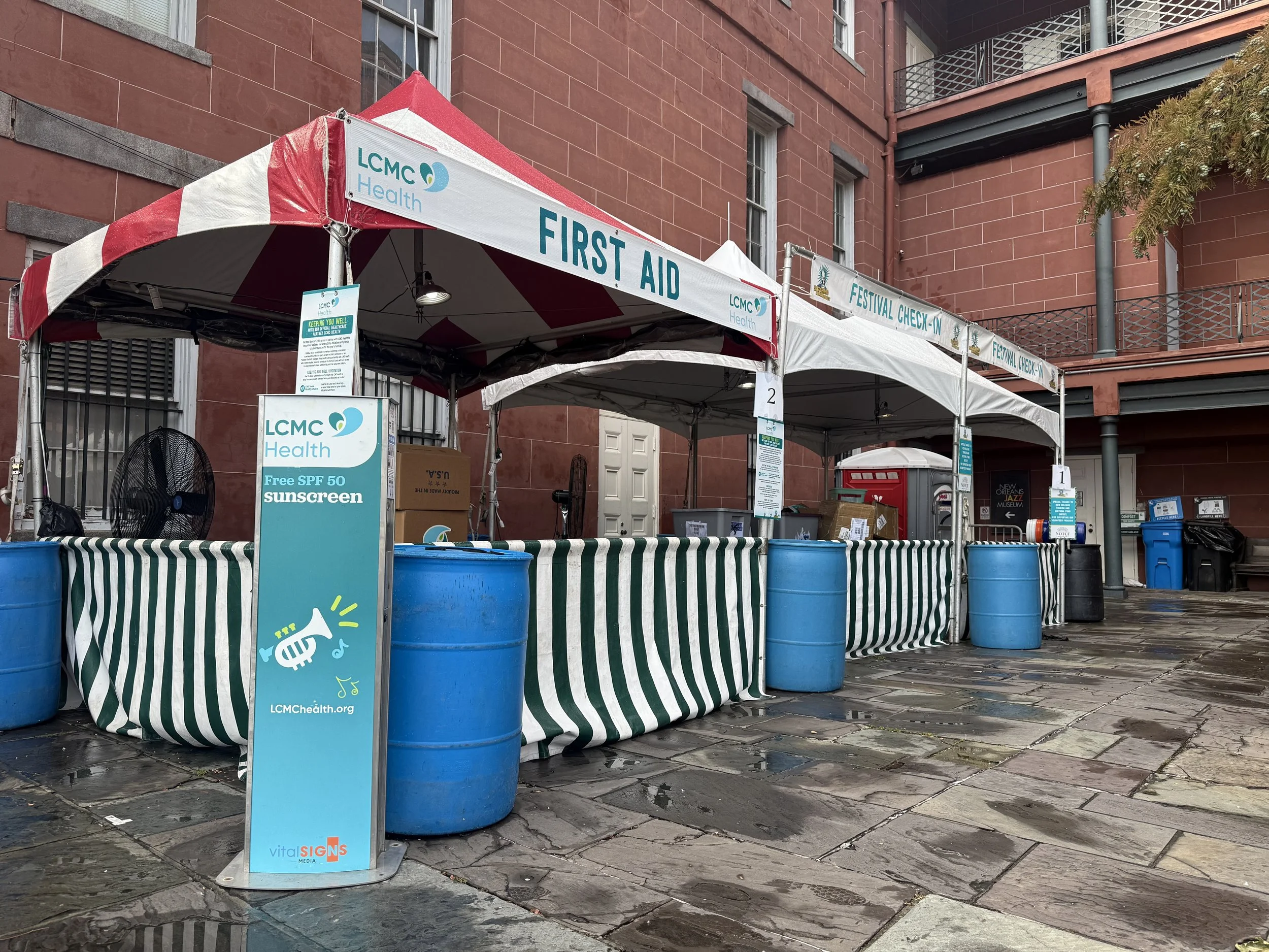 Outdoor first aid and festival check-in tents with striped green and white tablecloths, blue barrels, and health signage, set against red brick buildings on a wet stone-paved ground.