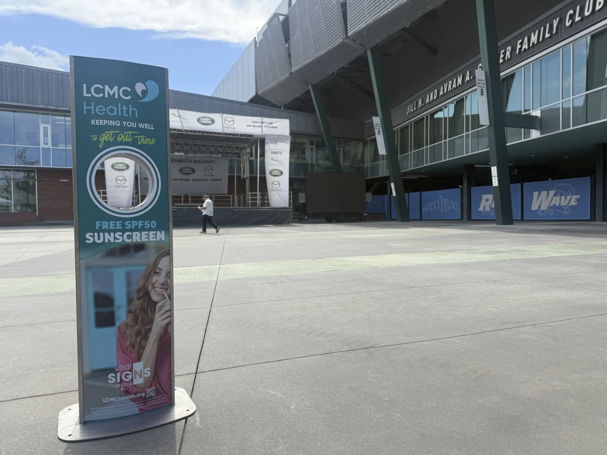 A kiosk sponsored by LCMC Health offering free SPF50 sunscreen outside Tulane's Yulman Stadium. The sign features a smiling woman and health-related branding.