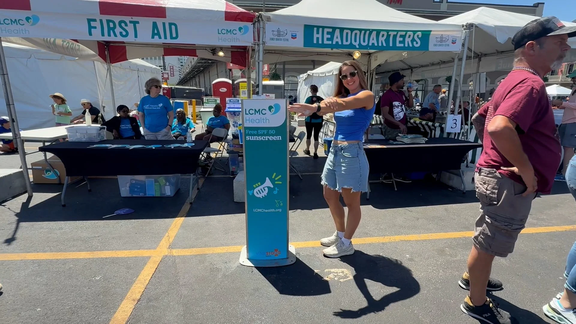 A woman smiling and putting on free SPF 50 sunscreen at a LCMC Health sponsored kiosk at an outdoor fair.