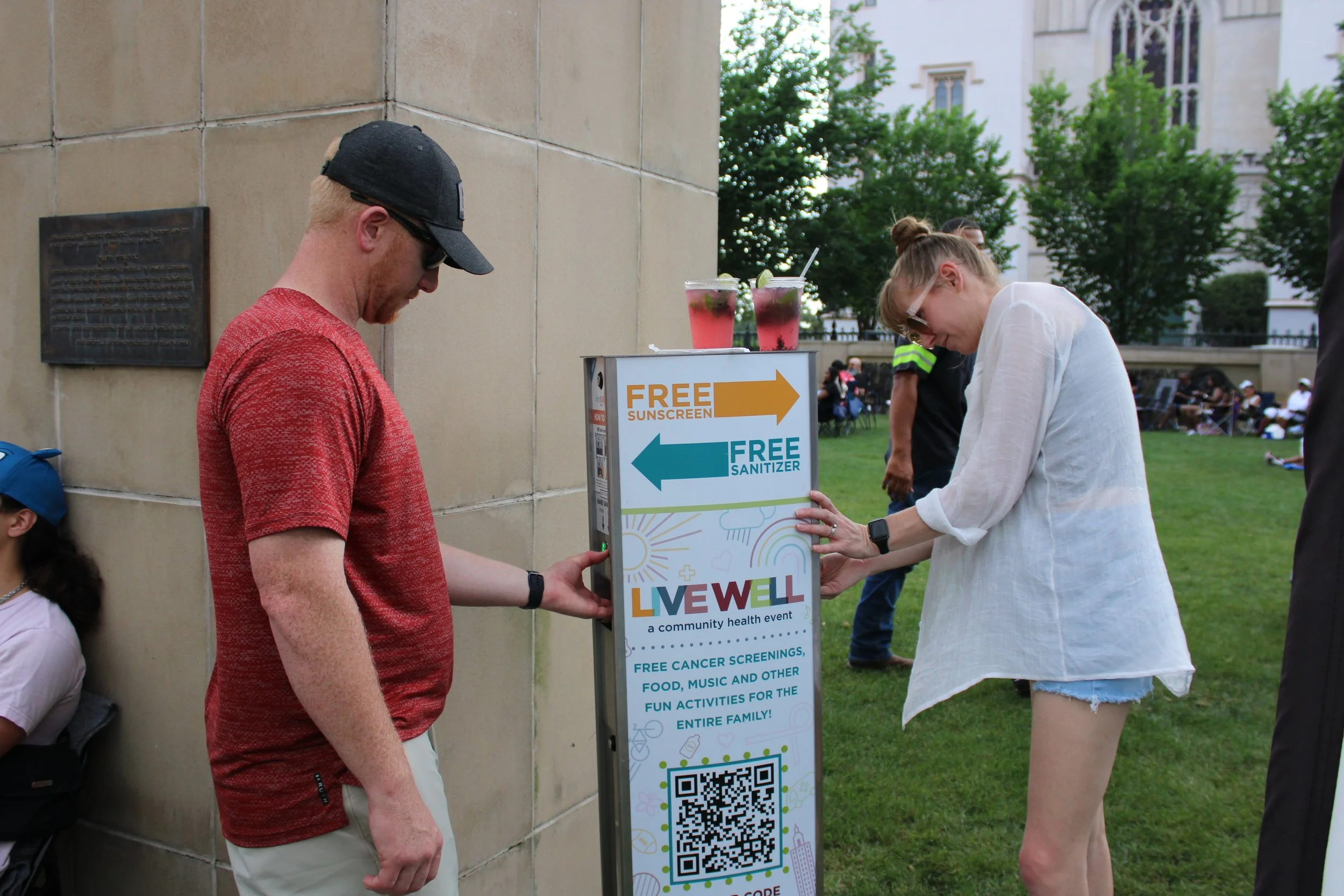 Two people by a Vital Signs Media sunscreen dispenser at a community health event offering free cancer screenings, food, music, and activities, with two drinks placed on top of the sign and a QR code for event details.
