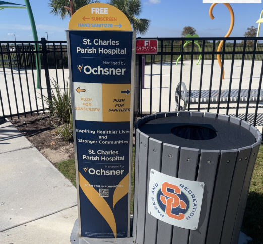 A public health and safety station with a sign advertising free sunscreen and hand sanitizer, next to a trash bin with USC Track & Recreation logo, in a sunny outdoor area near St. Charles Parish Hospital.
