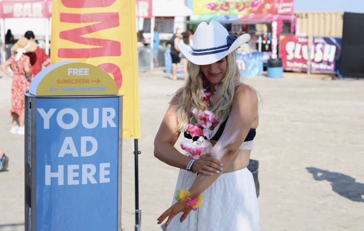 Woman on a beach dressed in a black bikini top, white skirt, and a white cowboy hat with a blue band, wearing flower leis and bracelets, standing near sunscreen dispenser saying, "Your Ad Here". 