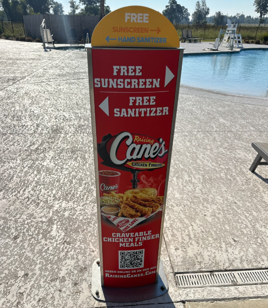 Kiosk at poolside offering free sunscreen and hand sanitizer, with Raising Cane's chicken fingers meal promotions and a QR code for online ordering.