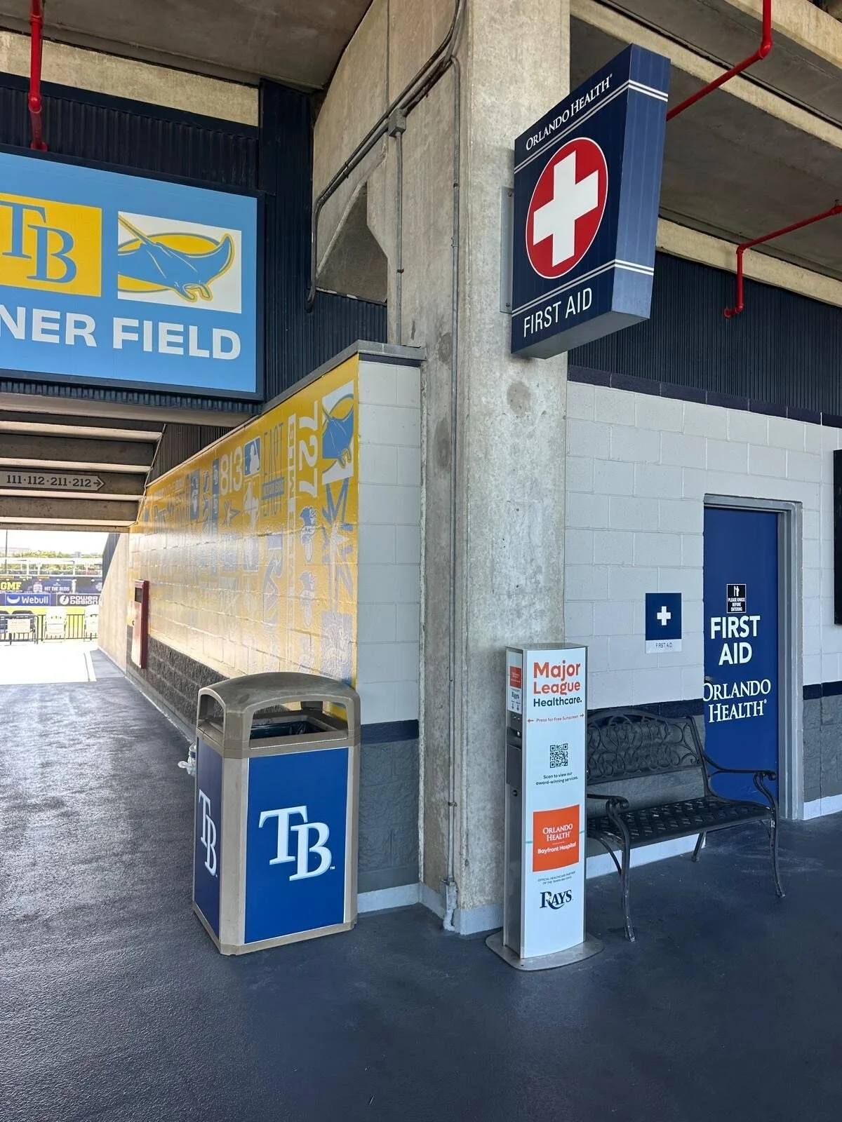 Sunscreen kiosk at the entrance to a sports stadium or event venue with TB and Major League Healthcare signs, a first aid sign, and a bench.