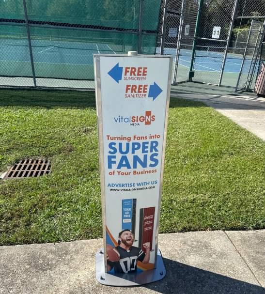 Outdoor sunscreen station advertising free sunscreen and sanitizer, with a URL for Vital Signs Media, and a promotional image of a person cheering holding a football and soda.