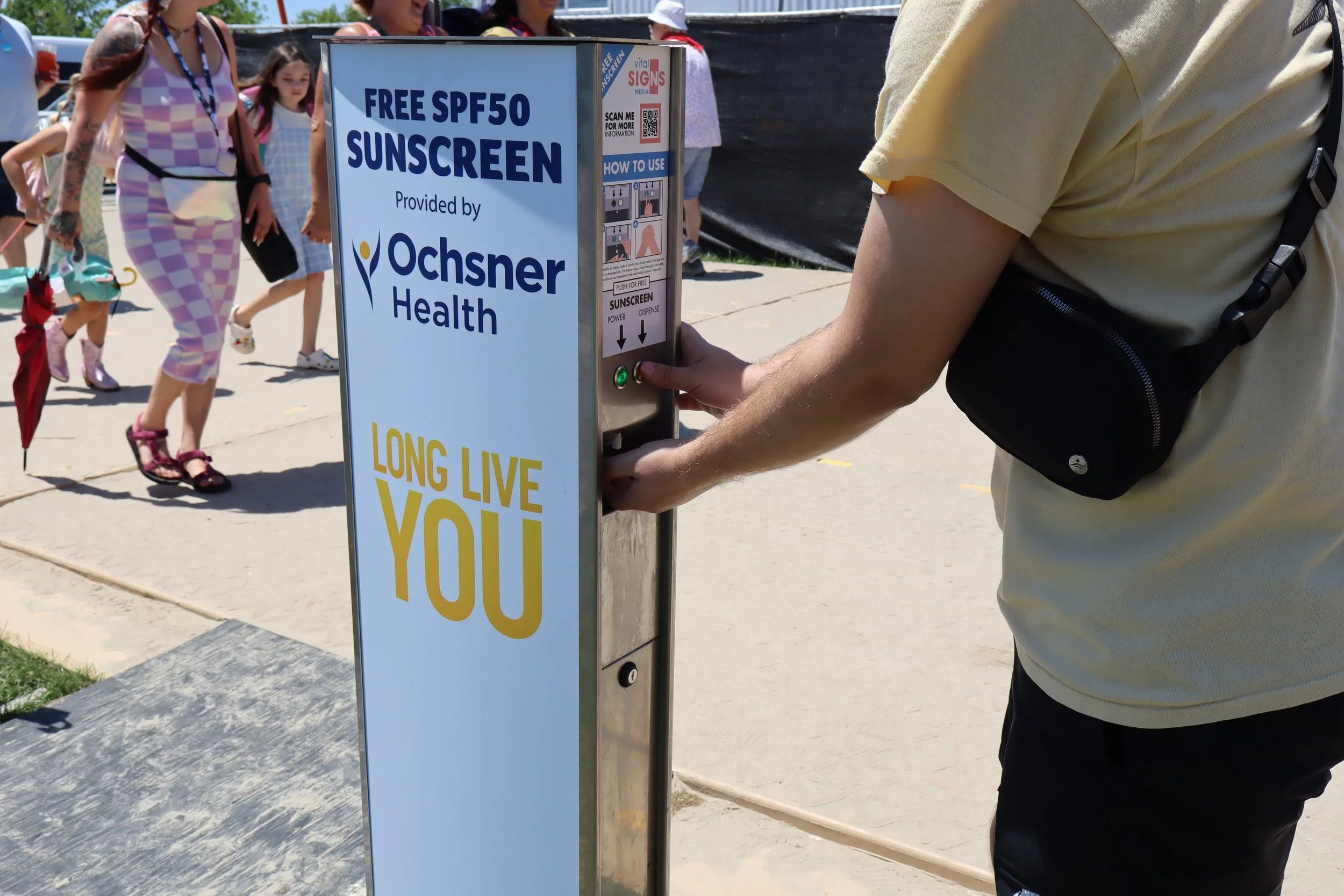 Person pressing button on a sunscreen dispenser with a sign advertising free SPF50 sunscreen provided by Ochsner Health, with children walking in the background at an outdoor event.
