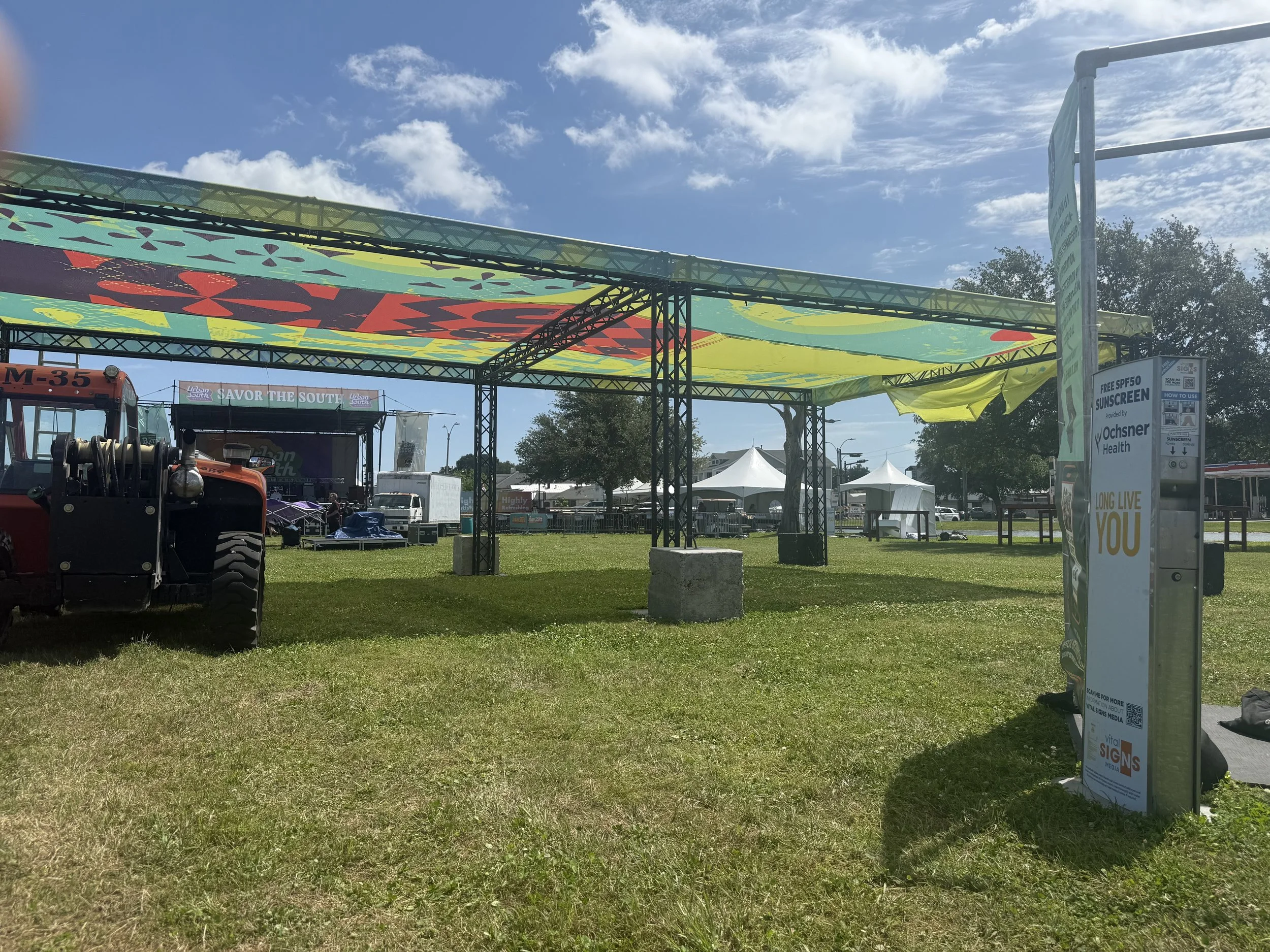 Open outdoor event area with a stage, tents, and a large colorful canopy, along with a fire truck parked on lush green grass and a bright blue sky with clouds.
