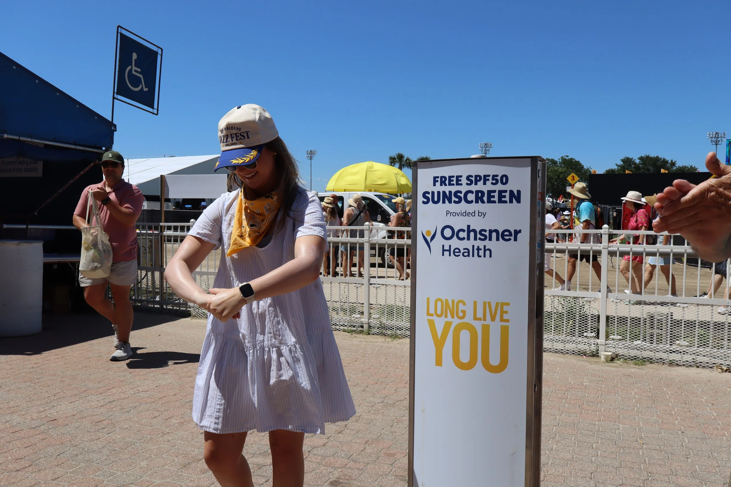 A woman wearing a white dress, a cap, and a yellow bandana smiling at a festival, with a sunscreen dispenser advertising free SPF50 sunscreen. There are people walking in the background at an outdoor event under a clear blue sky.