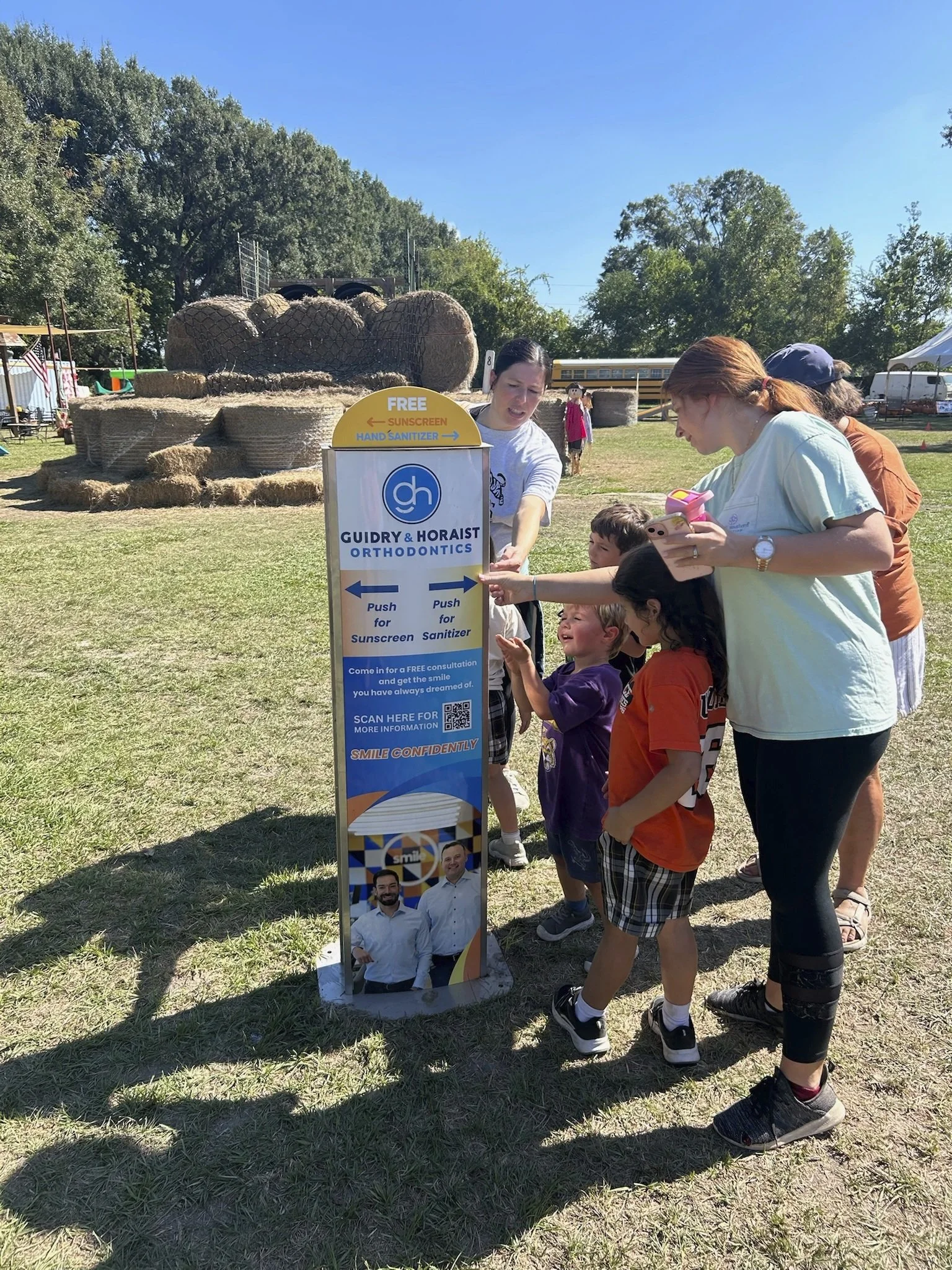 People gathered around a sign at an outdoor event, pointing and looking at the sunscreen kiosk which offers free sunscreen and hand sanitizer. The background features hay bales and a clear blue sky.