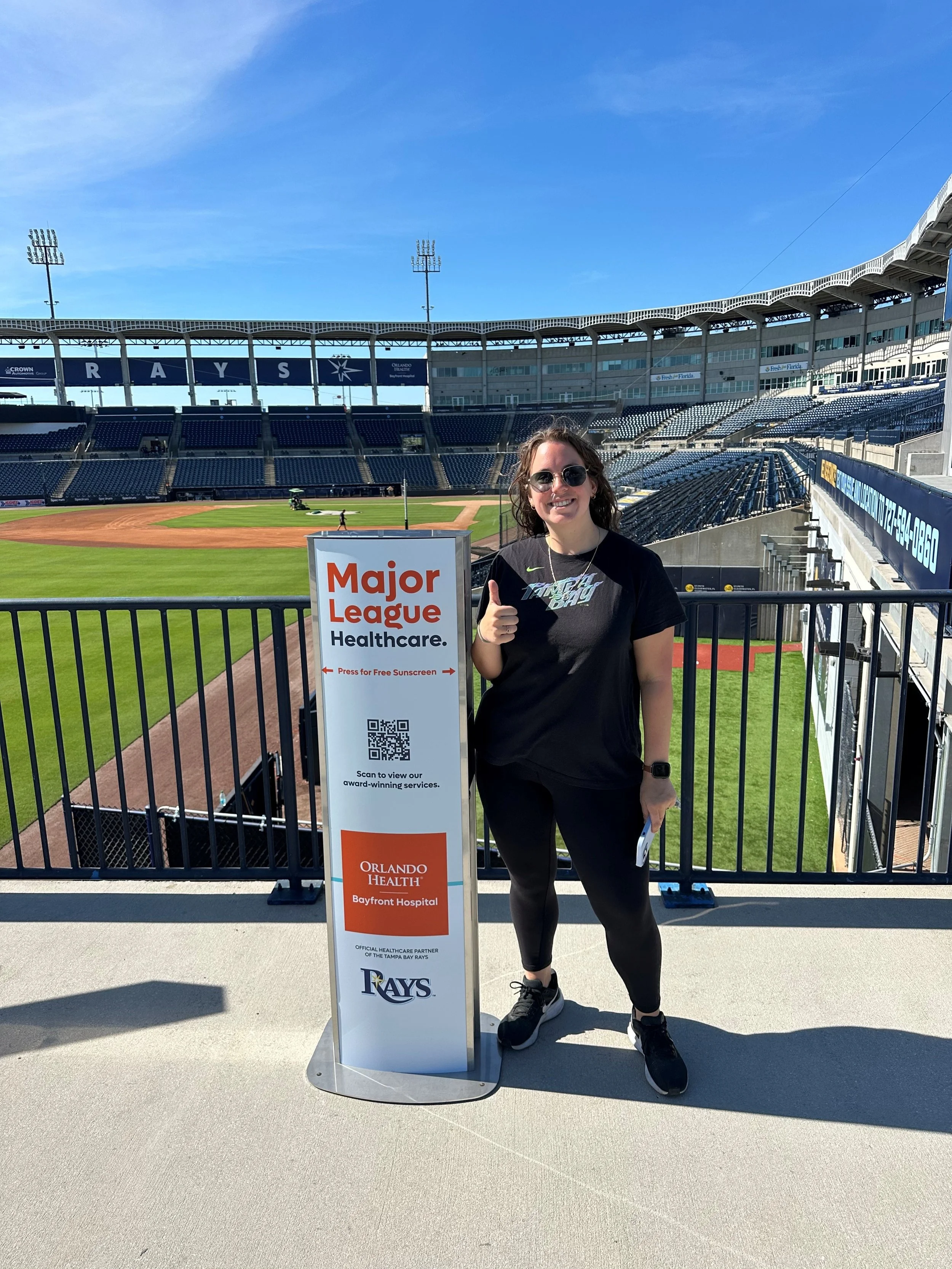 Woman in black athletic wear standing on stadium balcony with baseball field behind her, giving a thumbs-up, beside a Vital Signs media sunscreen dispenser sponsored by Major League Healthcare with a QR code, at a baseball stadium.